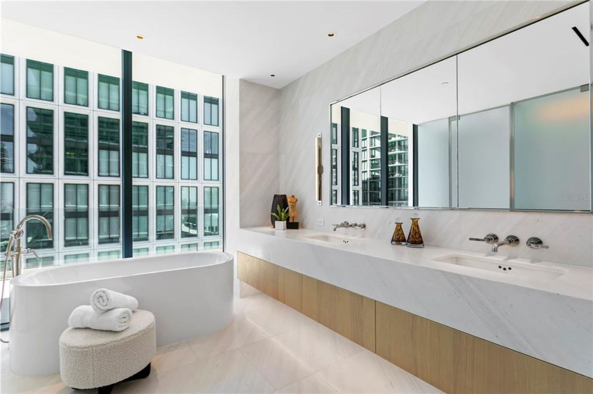 freestanding white bathtub near large floor-to-ceiling windows overlooking an urban view with buildings. A walk-in shower with a glass door and wall-mounted showerhead is located to the left. A small ottoman with neatly folded towels is placed next to the bathtub.