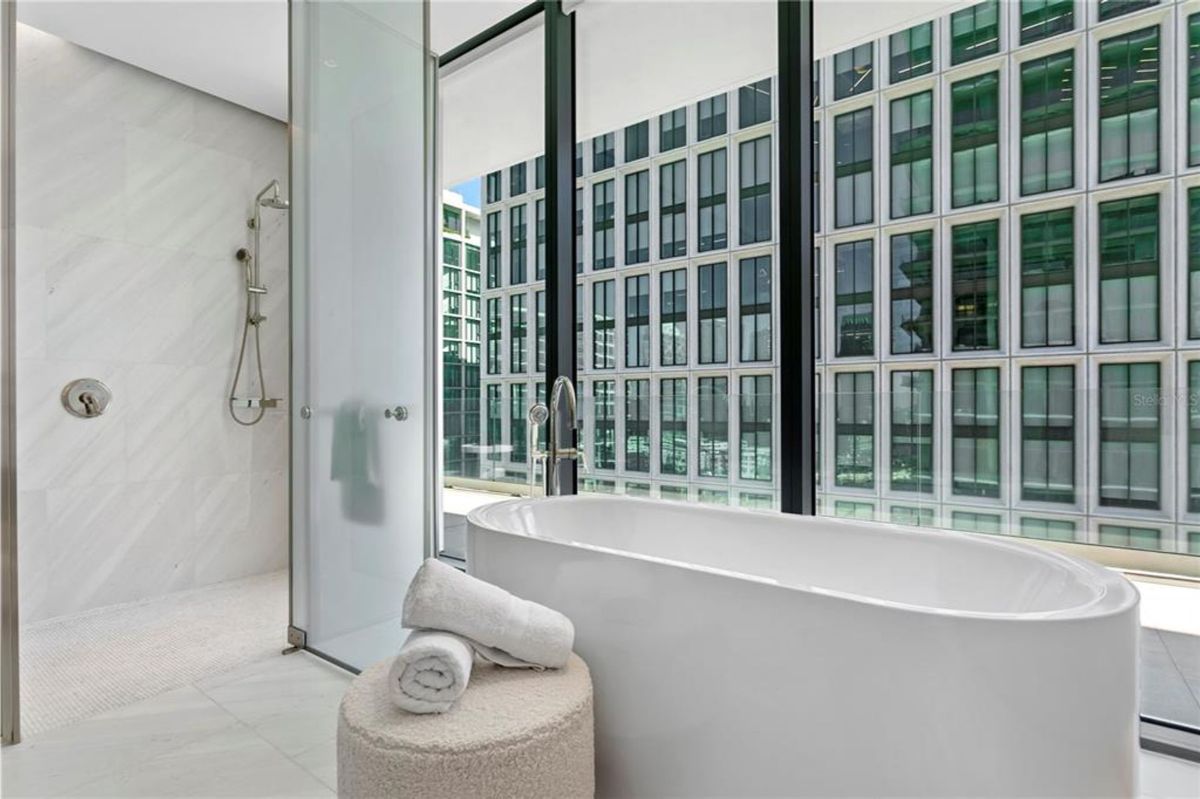 Bathroom with a freestanding white bathtub near large floor-to-ceiling windows overlooking an urban view with buildings. A walk-in shower with a glass door and wall-mounted showerhead is located to the left. A small ottoman with neatly folded towels is placed next to the bathtub.