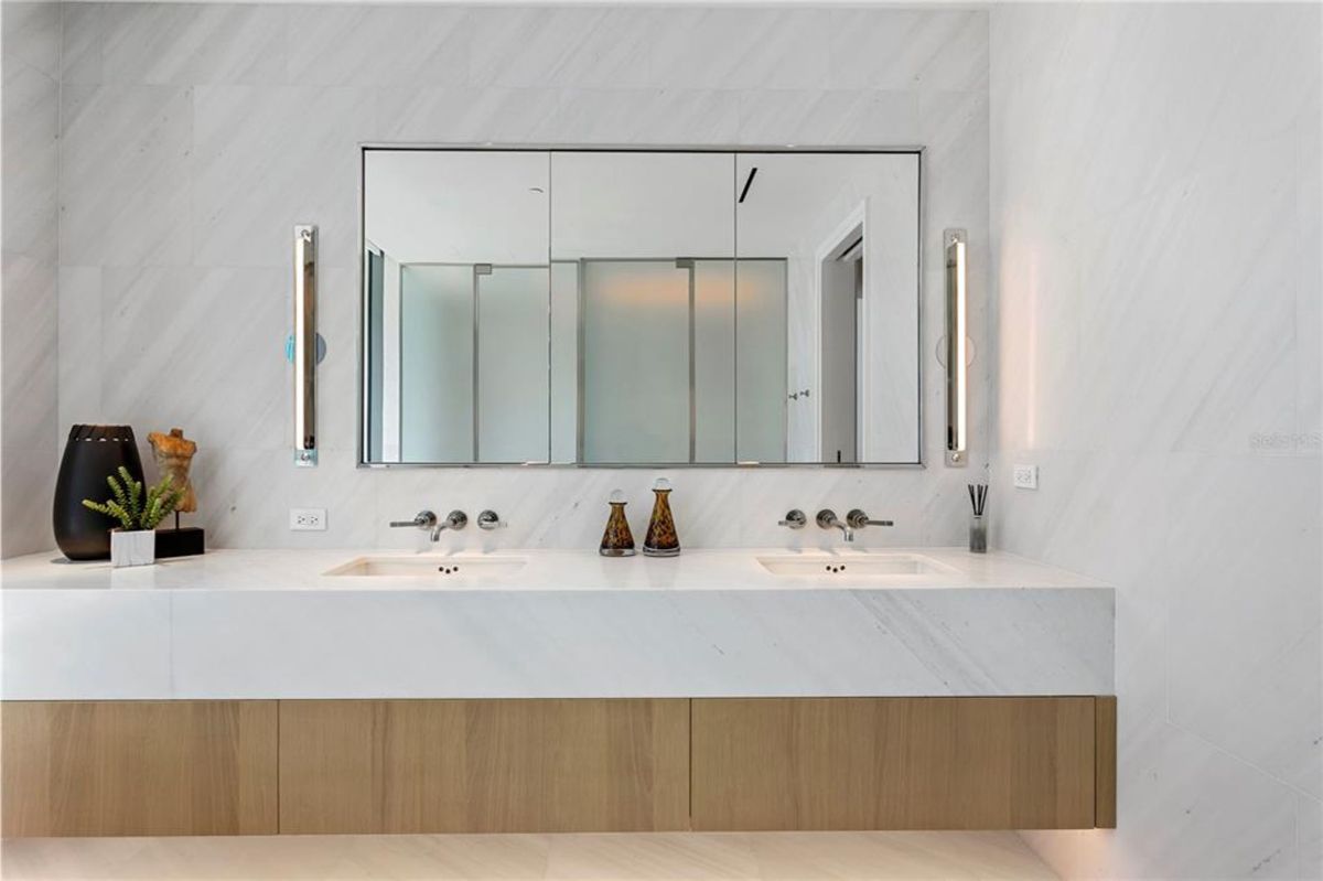 Double-sink vanity with a white marble countertop and a wooden cabinet base. Above the sinks, there is a large mirrored cabinet with three sections, flanked by two vertical wall-mounted lights. The space includes minimal decor such as a plant, small sculpture, and bottles, with walls covered in large light-colored tiles.