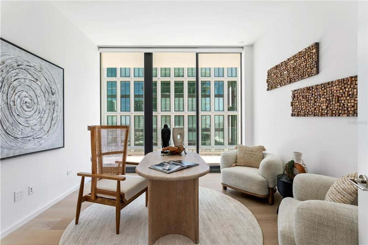 Sitting area with a wooden desk placed on a round textured rug. A high-backed wooden chair with woven elements is positioned on one side of the desk, while two armchairs with cushions and decorative accents are on the other side. Floor-to-ceiling windows offer views of a neighboring glass building, and the walls feature abstract art and wooden wall decorations.