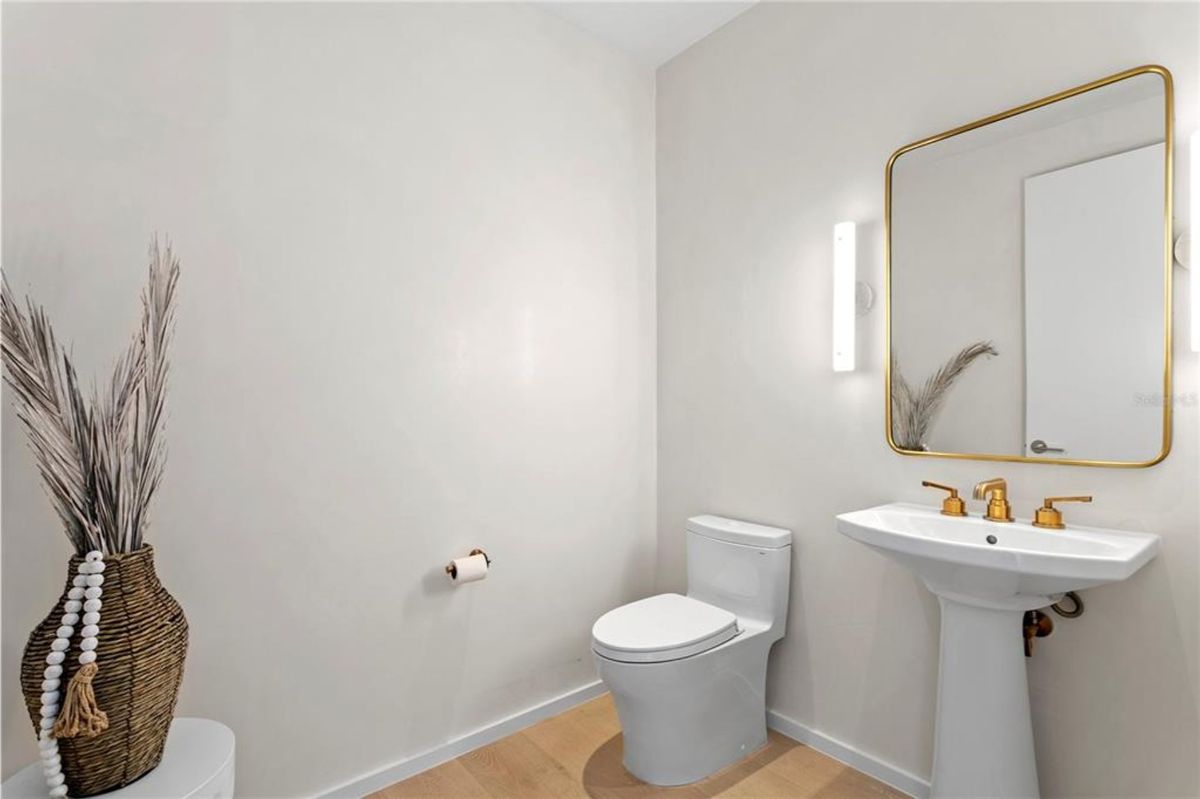 Bathroom with a white pedestal sink and a toilet. Above the sink, there is a rectangular mirror with a gold frame, flanked by two vertical wall lights. A decorative vase with tall dried plants is placed in the corner of the room, and a toilet paper holder is mounted on the wall.