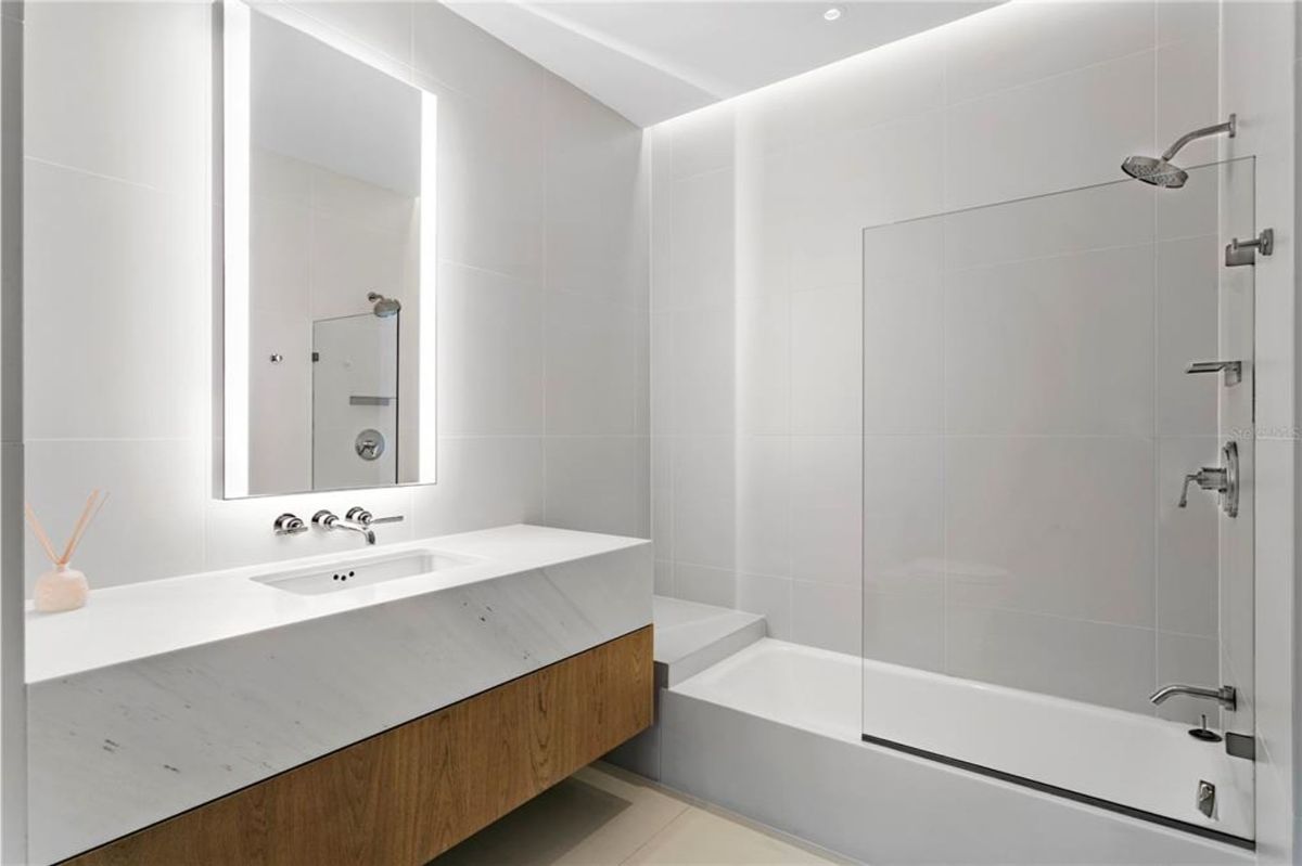 Bathroom with a white bathtub on the right, featuring a glass partition and a wall-mounted showerhead. On the left, there is a floating vanity with a white marble countertop, a rectangular sink, and a wooden cabinet below. Above the vanity, a large illuminated mirror is mounted on the wall, reflecting part of the bathroom.