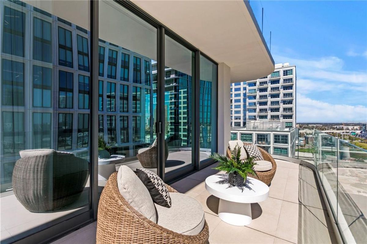 Balcony with glass railings and a view of nearby modern high-rise buildings. The space has two wicker chairs with beige cushions, a small white round table, and a potted plant on the table. The sliding glass doors lead into the adjacent interior, reflecting the outdoor environment.