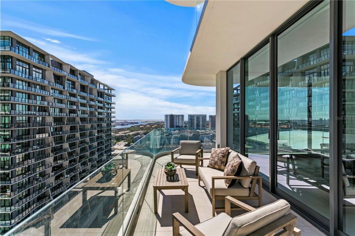 Balcony with glass railings and a view of surrounding high-rise buildings and cityscape. There are outdoor wooden furniture pieces, including two chairs, a loveseat, and a coffee table, with cushions and throw pillows. The balcony connects to the interior through large glass sliding doors, which reflect the city skyline.