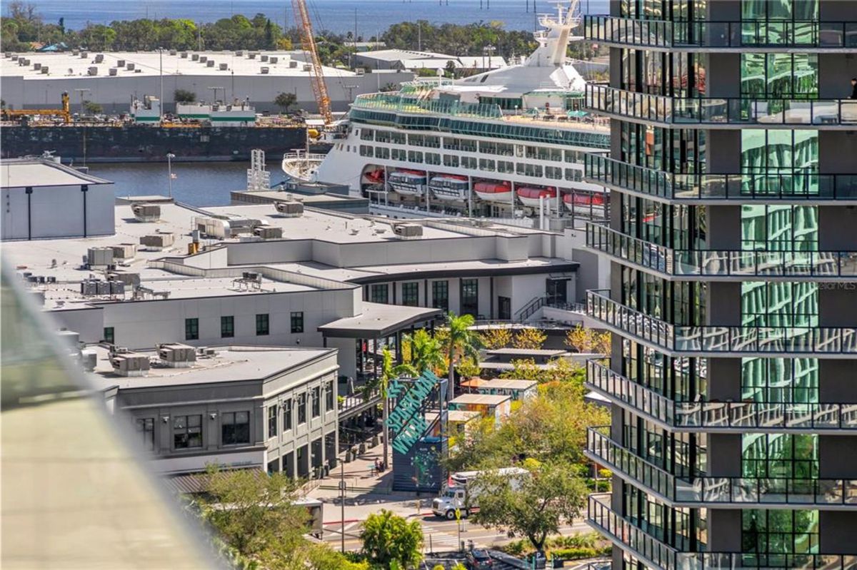 A large docked cruise ship next to a port area with industrial and commercial buildings in the foreground. A nearby high-rise building with reflective glass windows dominates the right side of the frame. Visible signage, streets, and cranes suggest the location is part of a busy waterfront city or cruise terminal.