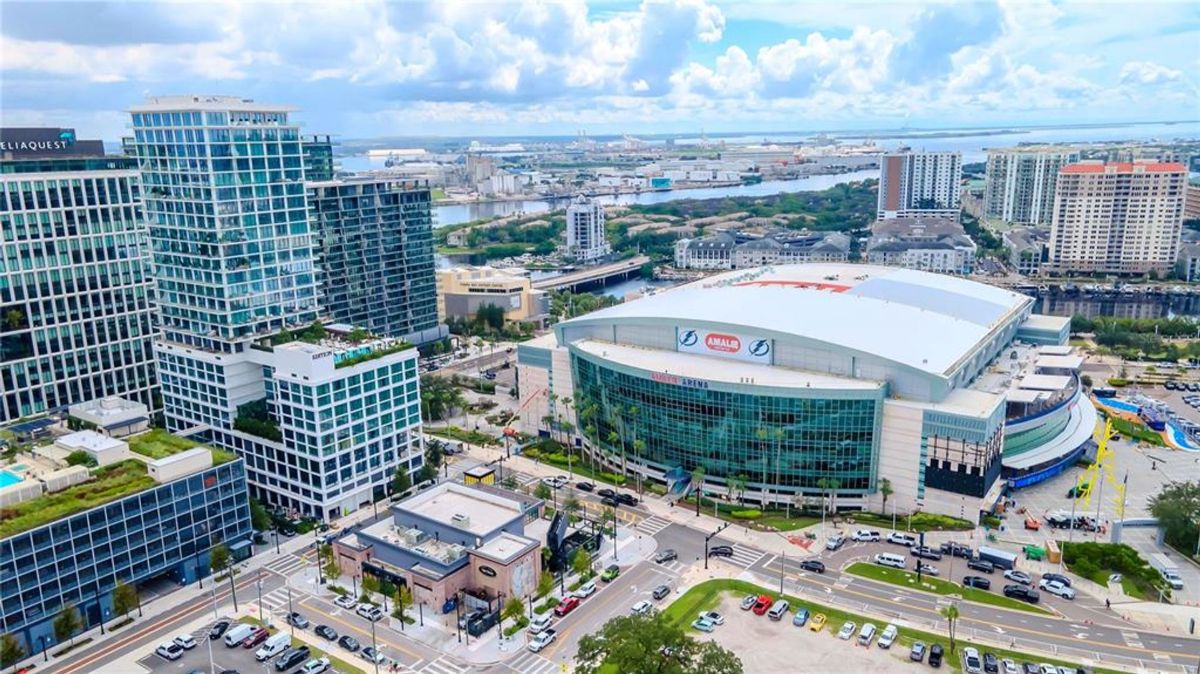 A large urban area with modern high-rise buildings and an arena labeled "Amalie Arena" at the center. The arena has a curved roof and large glass windows on its front, surrounded by roads, parked cars, and nearby structures. In the background, there is a waterfront area with additional buildings, water, and industrial activity visible.