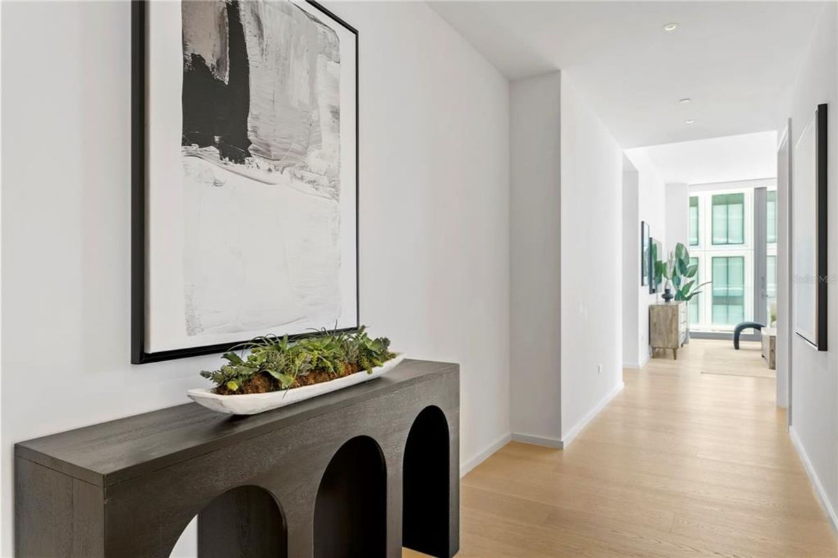 Hallway with a large abstract painting in black and white on the left wall. Below the painting, a black console table with arched cutouts holds a decorative planter filled with greenery. At the end of the hallway, a room with floor-to-ceiling windows, a cabinet, and a large indoor plant is visible.