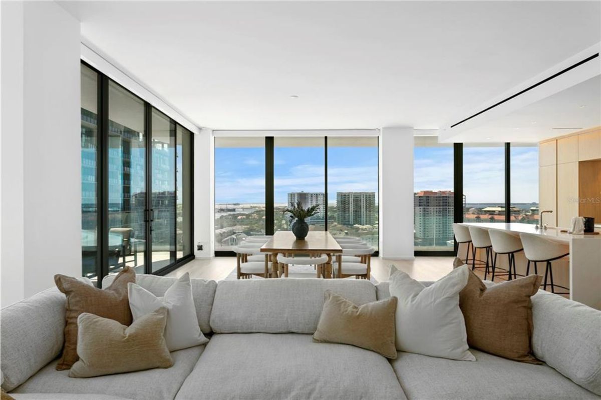 Open living space with a large light gray sectional sofa adorned with multiple throw pillows. In the background, a dining table with chairs is set near floor-to-ceiling windows offering views of nearby buildings and the horizon. To the right, a kitchen area with a counter, barstools, and cabinets is visible, integrating seamlessly with the rest of the space.