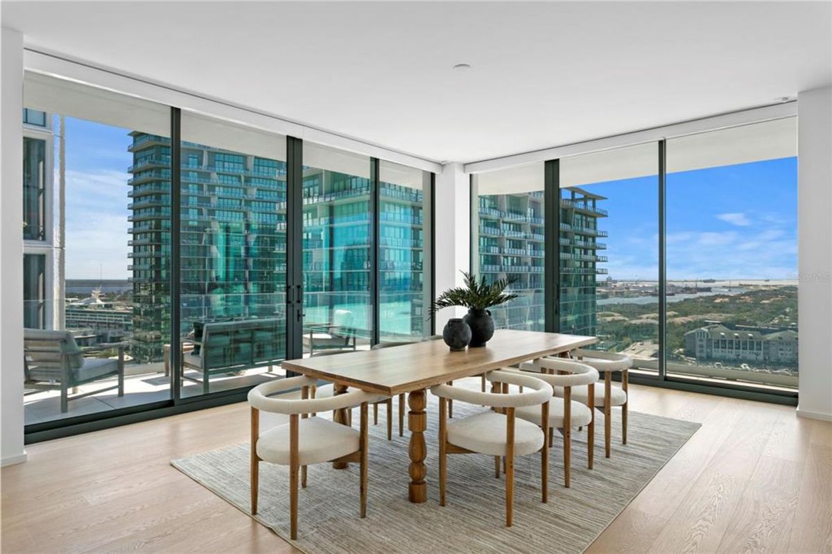 Dining room with a rectangular wooden table and six upholstered chairs on a textured rug. Large floor-to-ceiling glass windows surround the space, offering views of nearby glass buildings and a distant landscape. Two decorative vases with green plants are placed as a centerpiece on the table.