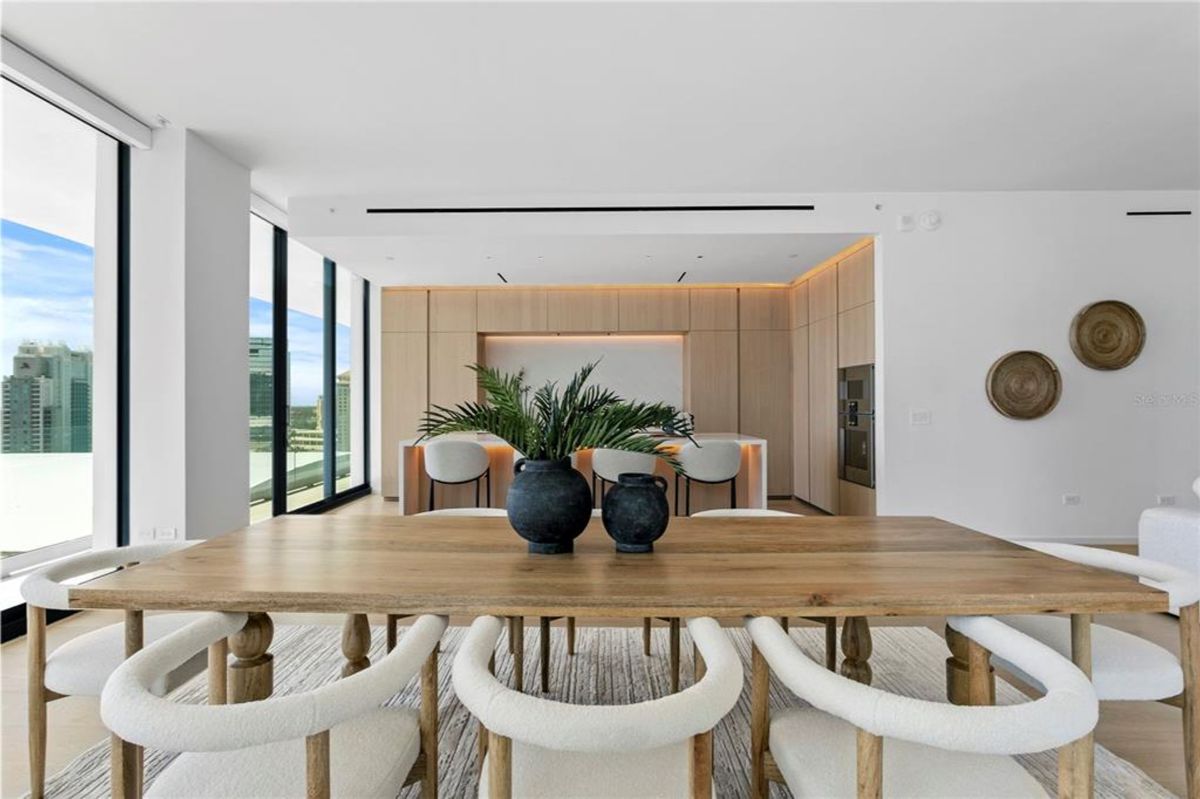 A wooden dining table with eight white upholstered chairs, placed on a textured rug in an open-concept dining area. Two black decorative vases with plants are centered on the table. In the background, there is a kitchen with wood-paneled cabinetry, a white countertop island, and three white bar stools, along with two decorative wall pieces on the right.