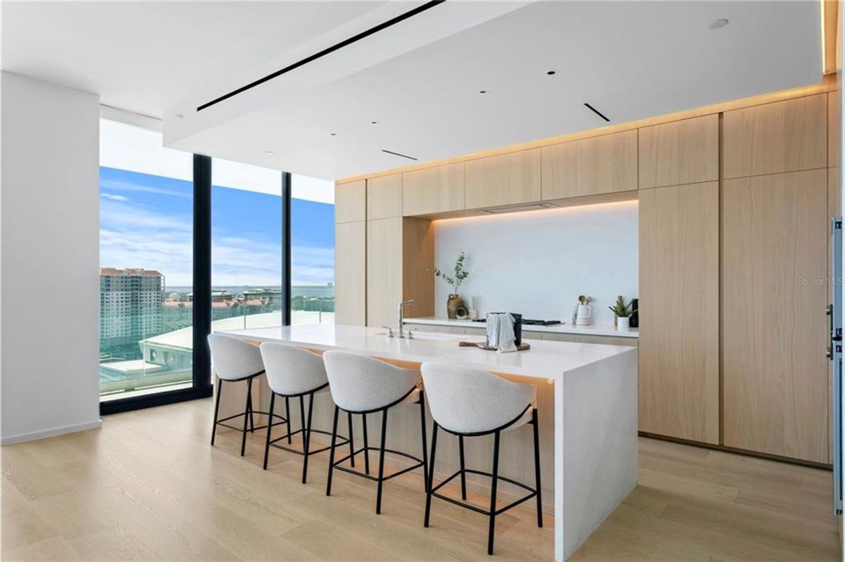 Kitchen with a large white island countertop that includes a built-in sink and four upholstered bar stools. Behind the island is a wall of light wood cabinetry with integrated appliances and a backsplash with soft lighting. Floor-to-ceiling windows provide natural light and offer views of nearby buildings and the cityscape.