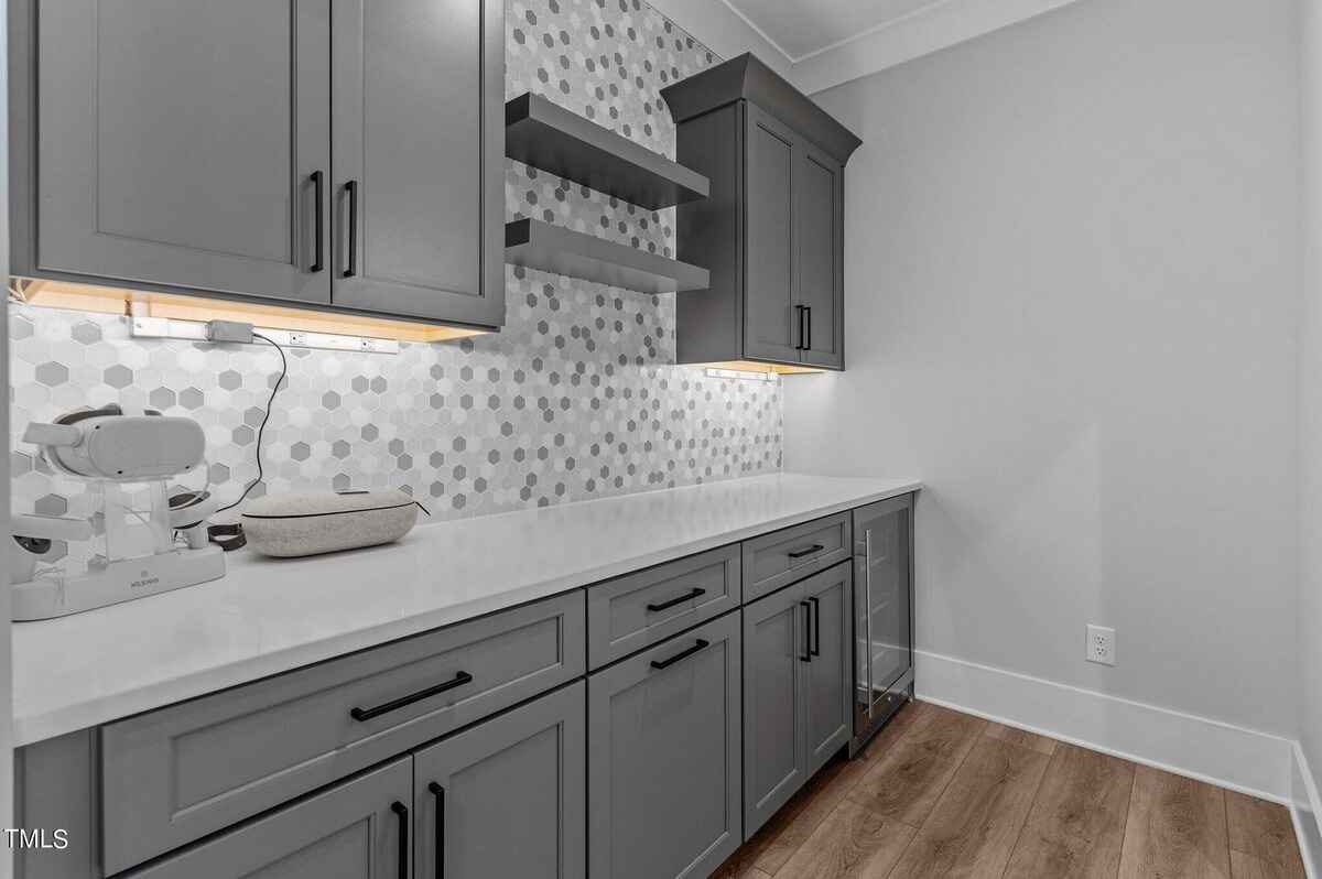 Gray cabinets and white countertops line a wall with hexagonal tile backsplash and floating shelves.