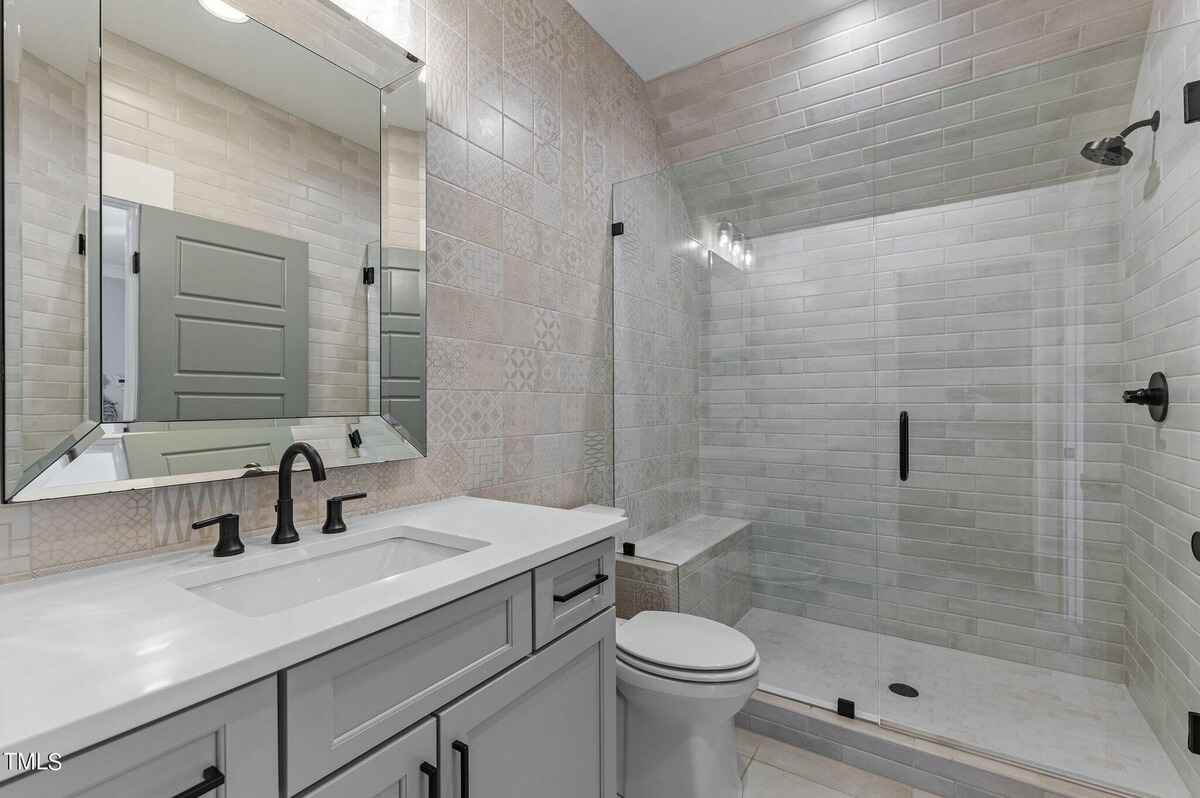 Bathroom features gray vanity with white countertop and black hardware, glass shower with built-in bench, and patterned tile walls.