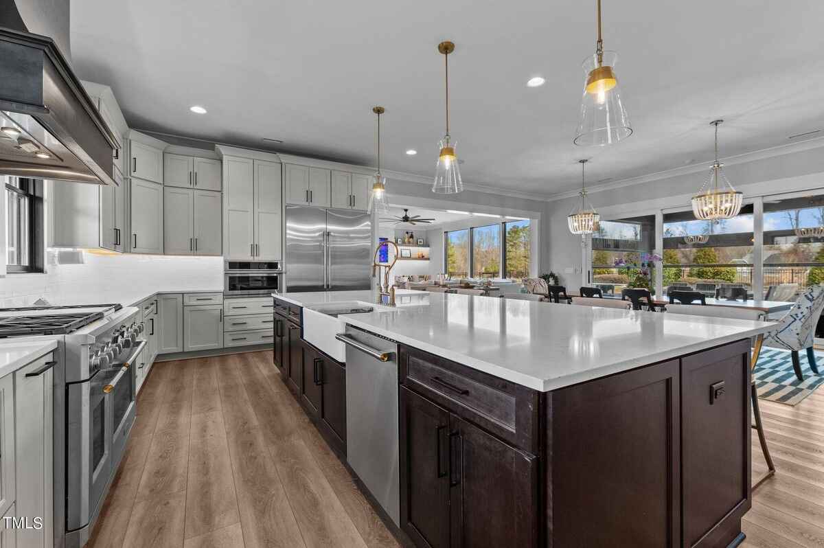 Kitchen boasts light gray cabinets, dark brown island with a farmhouse sink, stainless steel appliances, and pendant lighting, overlooking a dining and living area.