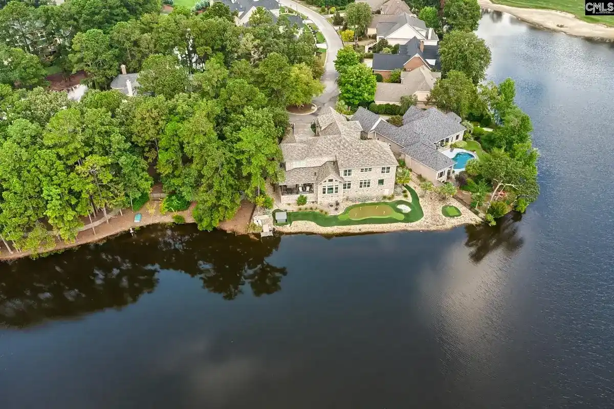 An aerial shot shows a large house situated on the edge of a lake, surrounded by lush greenery and other houses.