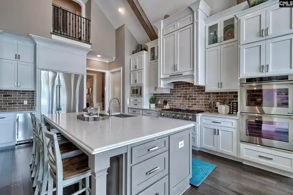 A kitchen with white cabinets, gray island, and a brick backsplash features a large island with seating and high ceilings.