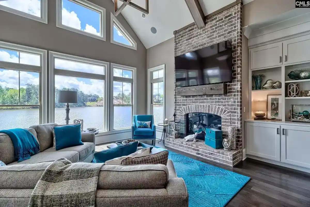 A living room with vaulted ceilings, large windows overlooking a lake, a brick fireplace with a mounted TV, and a teal and gray color scheme is shown.