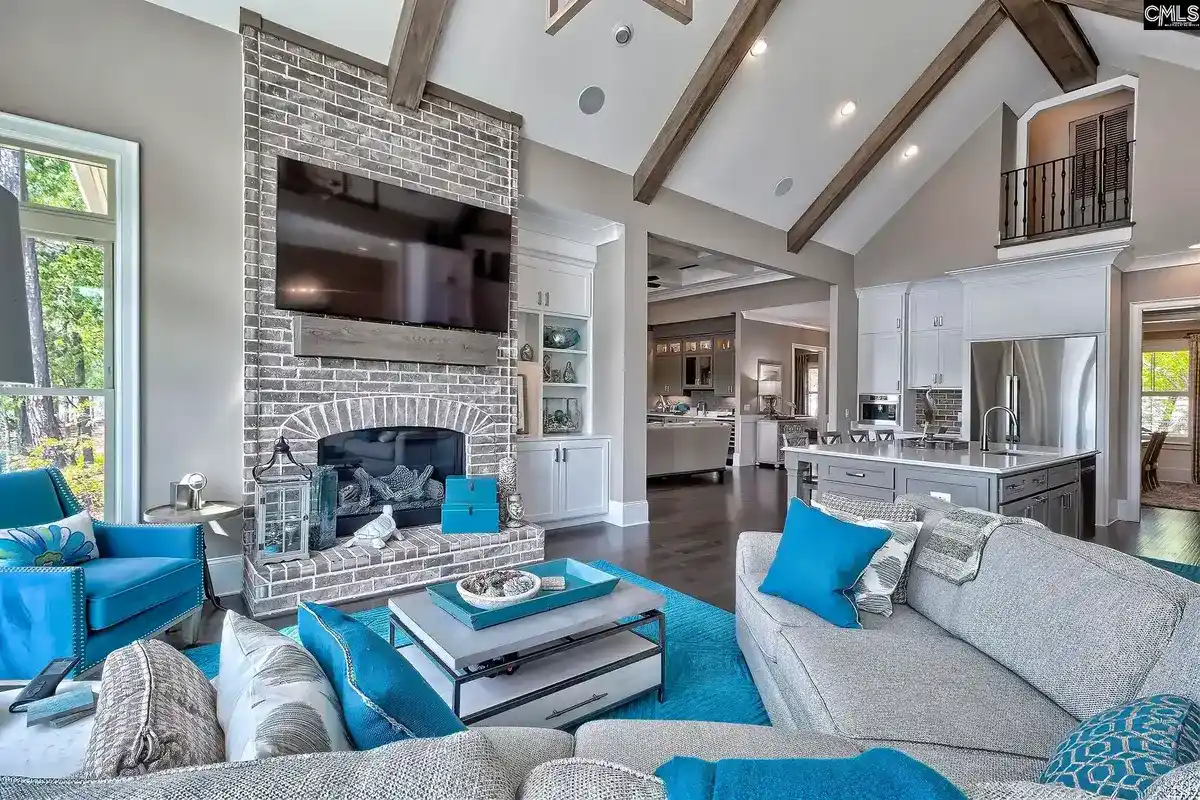 A spacious living room with high ceilings, exposed beams, a large fireplace, and a teal and gray color scheme opens to a kitchen and dining area.