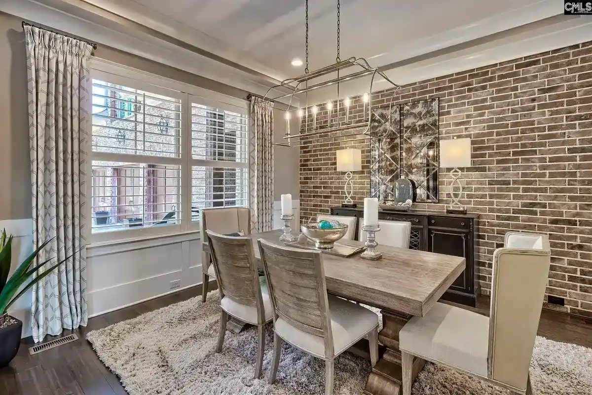 A formal dining room features a long wooden table surrounded by light-colored chairs, a brick accent wall, and a large chandelier.