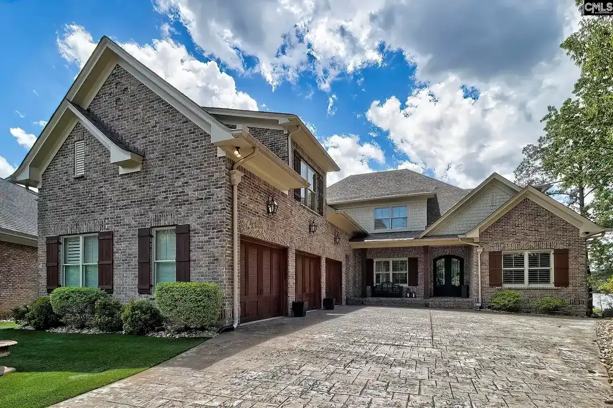 A large brick house with a three-car garage and a stamped concrete driveway sits under a partly cloudy blue sky.