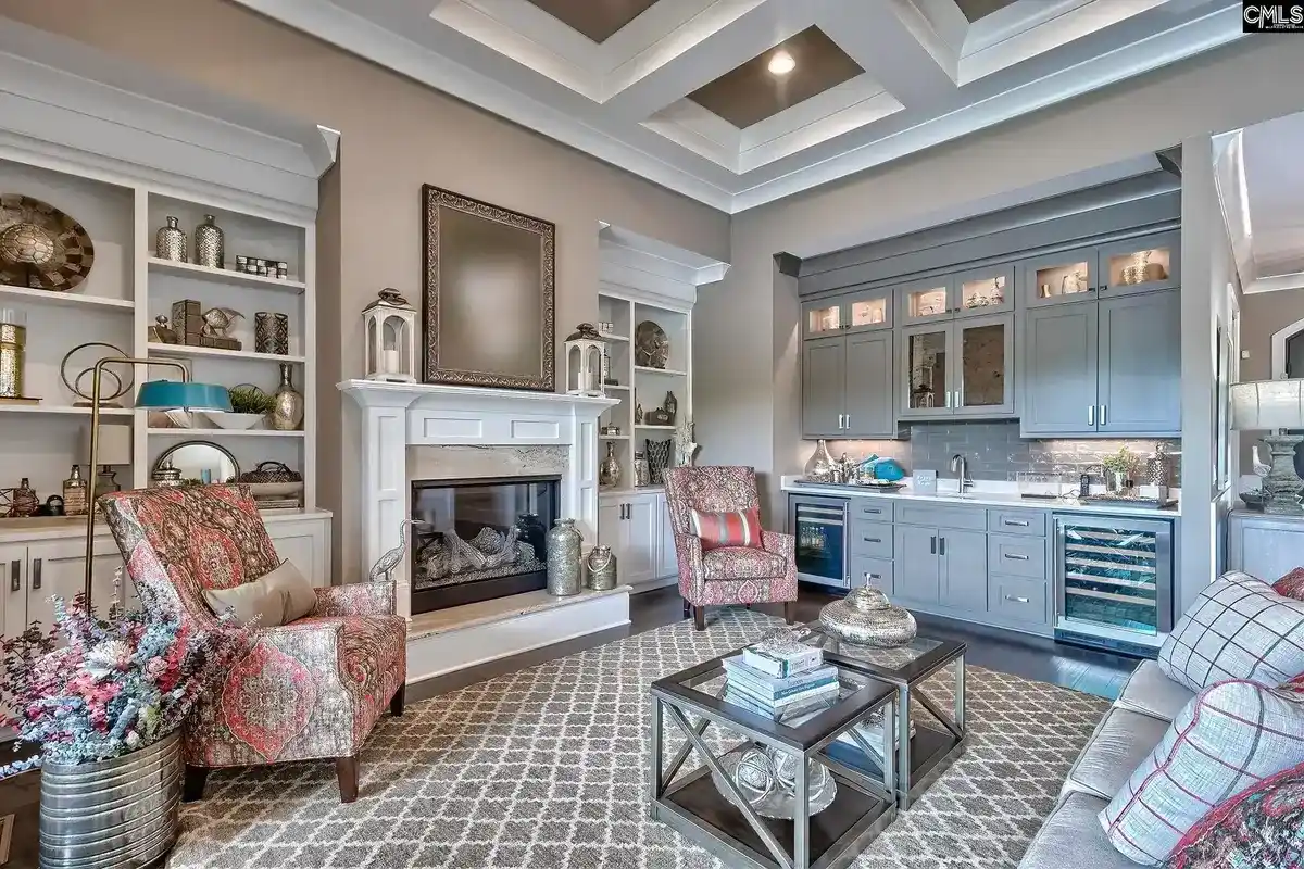 A cozy sitting area with built-in bookshelves and a fireplace is adjacent to a wet bar with gray cabinets and a wine refrigerator.