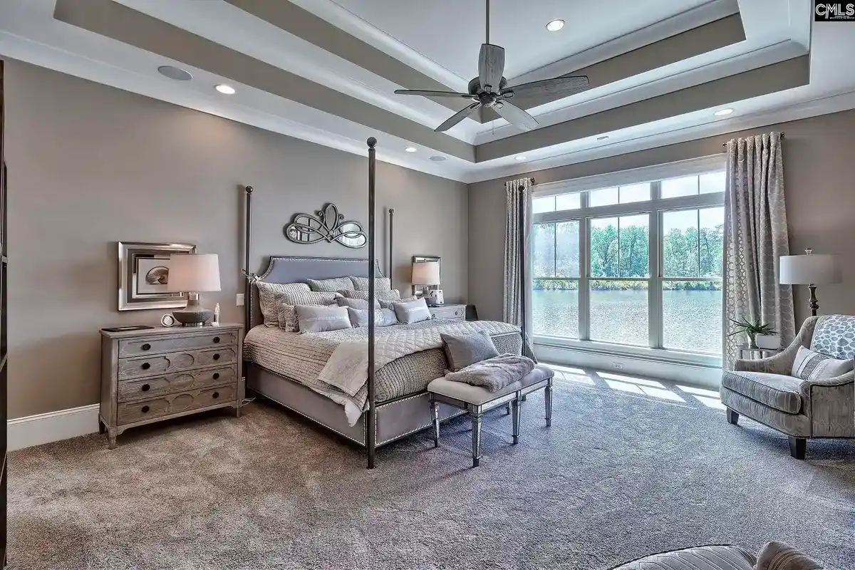 A spacious master bedroom with a four-poster bed, large windows offering a lake view, and a neutral color palette is shown.