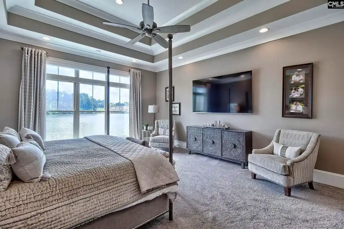 A master bedroom with a four-poster bed, a large flat-screen TV, and a ceiling fan features a sitting area with a chair and a view of a lake from the window.