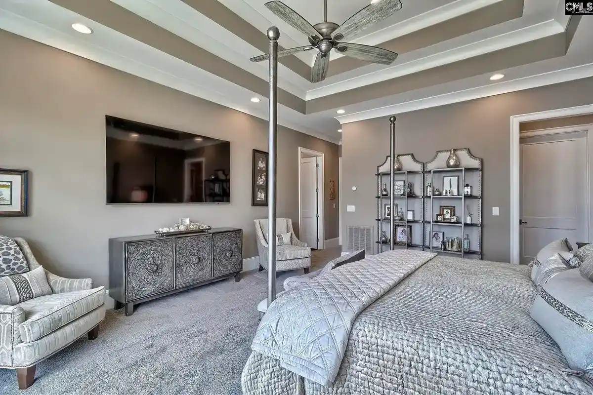 A large master bedroom with a four-poster bed, a flat-screen TV, and a ceiling fan features a neutral color scheme and decorative furniture.