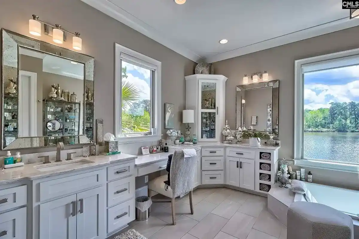 A spacious bathroom with white cabinets, double vanities, and a large soaking tub boasts a view of a lake through a large window.