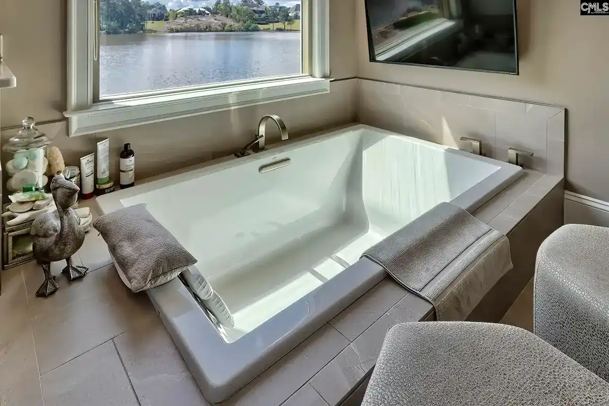 A luxurious soaking tub with a lake view is situated in a modern bathroom with neutral tones and a mounted television.