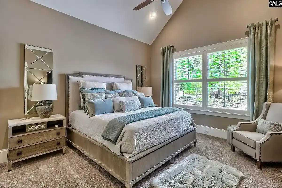 A bedroom with a large bed, numerous pillows, and a neutral color palette features a window with white shutters and a comfortable armchair.