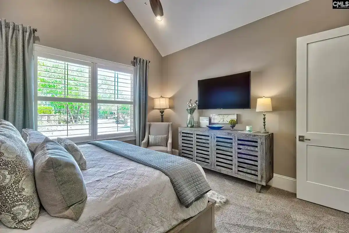 A bedroom with a large bed, a flat-screen TV, and a gray color scheme features a window with white shutters and a gray dresser.