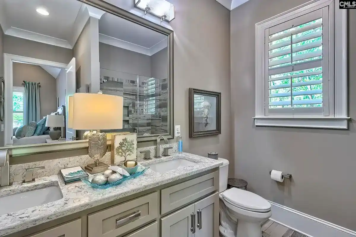 A bathroom with a double vanity, granite countertops, and a large mirror features a toilet and a window with white shutters.