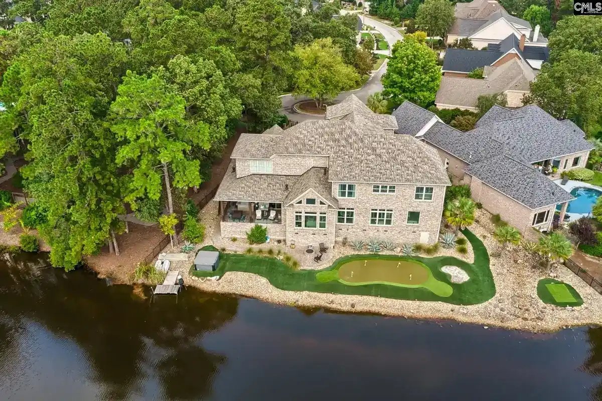 An aerial view shows a large house on a lake with a putting green in the backyard.