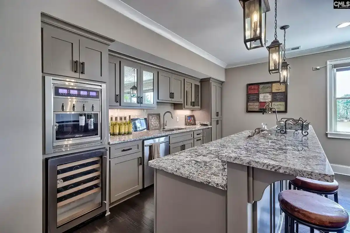 A kitchen with gray cabinets, granite countertops, and a wine refrigerator features an island with bar stools and pendant lighting.