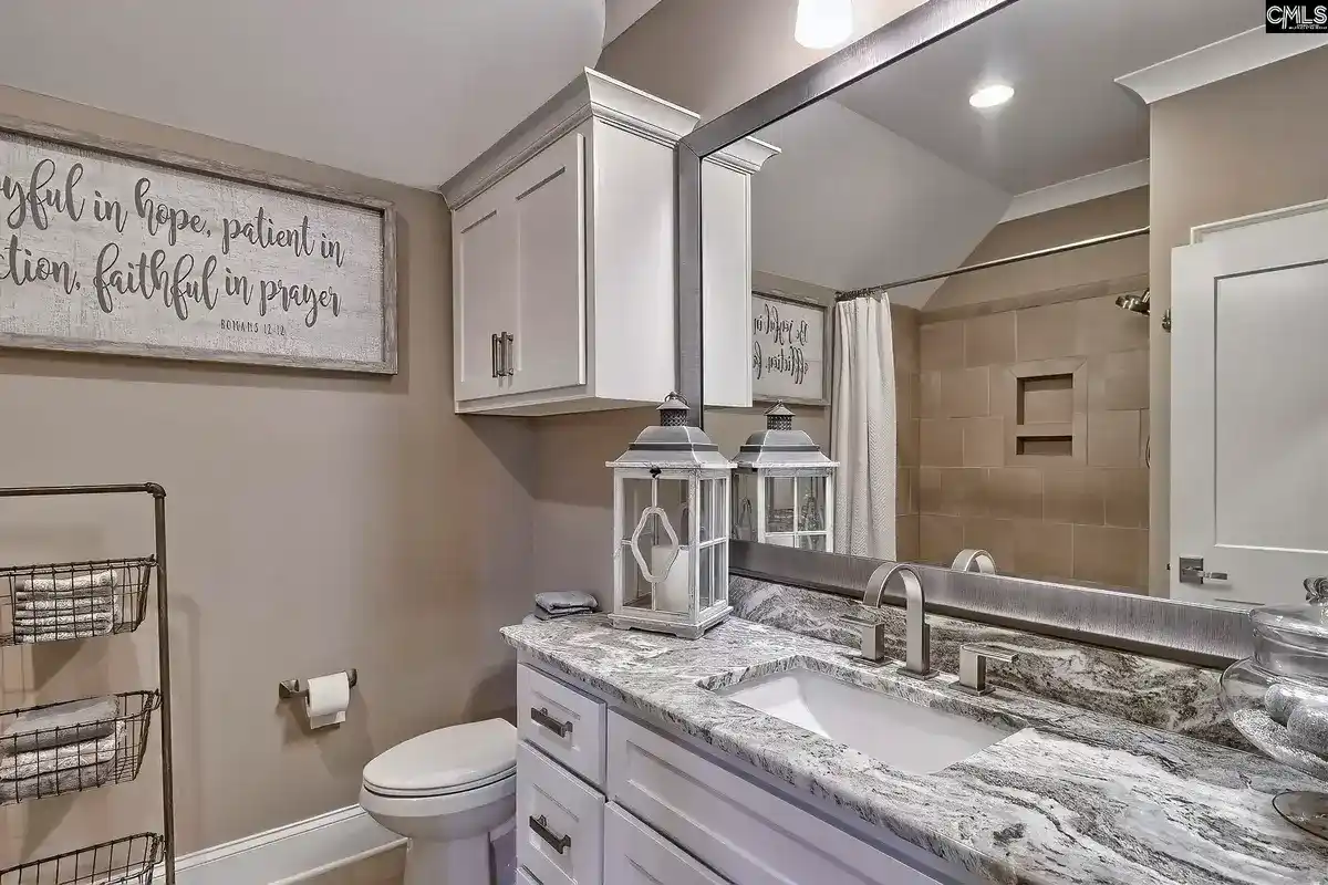 A bathroom with a vanity, granite countertop, and a large mirror features a framed Bible verse and a shower with beige tile.