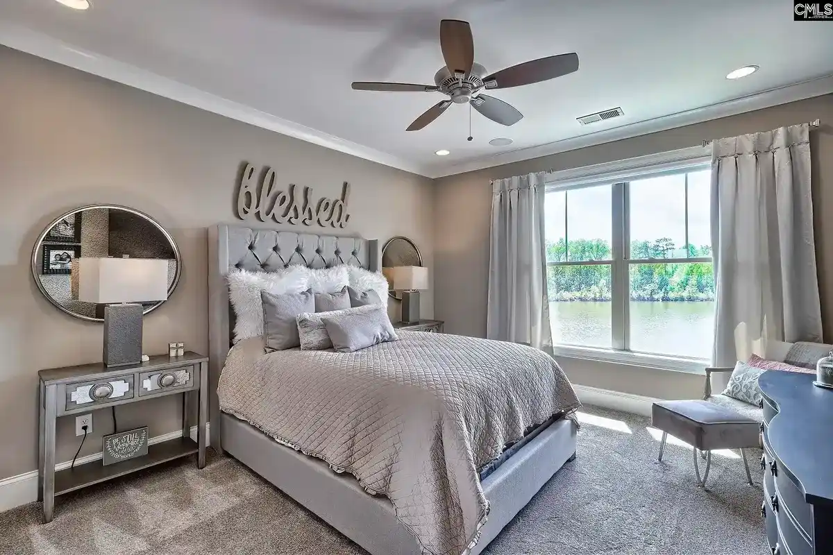 A bedroom with a neutral color scheme features a tufted headboard, a ceiling fan, and a window overlooking a lake.