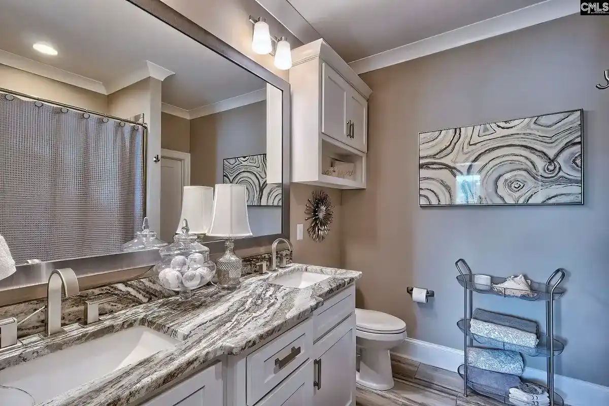 A bathroom with a large granite countertop, double sinks, and a neutral color scheme features a large mirror and artwork above a toilet.