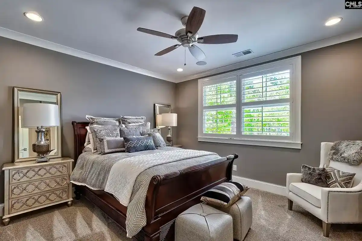 A bedroom with a dark wood sleigh bed, neutral-toned bedding, and a ceiling fan features a sitting area with a white armchair and a window with white shutters.