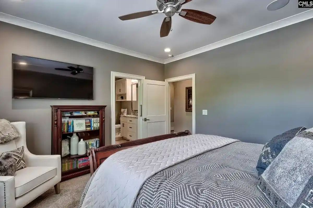 A bedroom with a ceiling fan, a large flat-screen TV, and a gray color scheme features a bed with a geometric patterned comforter and a white armchair.