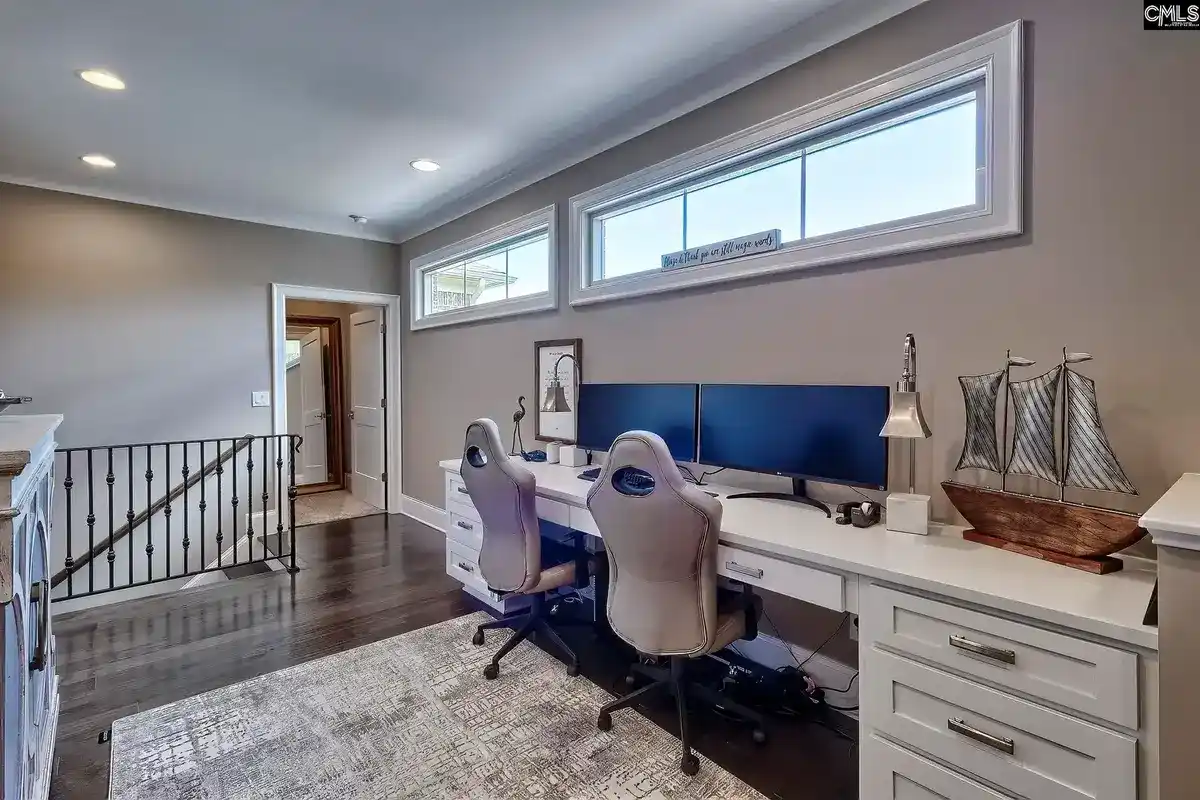 A home office with two computer workstations, a white desk, and built-in cabinets is situated near a staircase and features large windows.