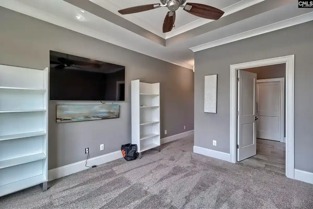 A spacious room with gray walls and carpeting features a large flat-screen TV mounted above a long piece of artwork, flanked by white bookshelves, and a ceiling fan.