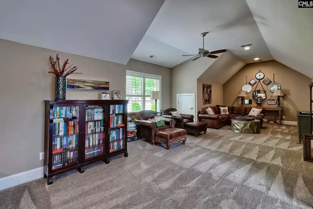 A large, carpeted room with a vaulted ceiling, a ceiling fan, and a brown leather sectional sofa features a bookcase filled with books and a wall decorated with a collection of mirrors.