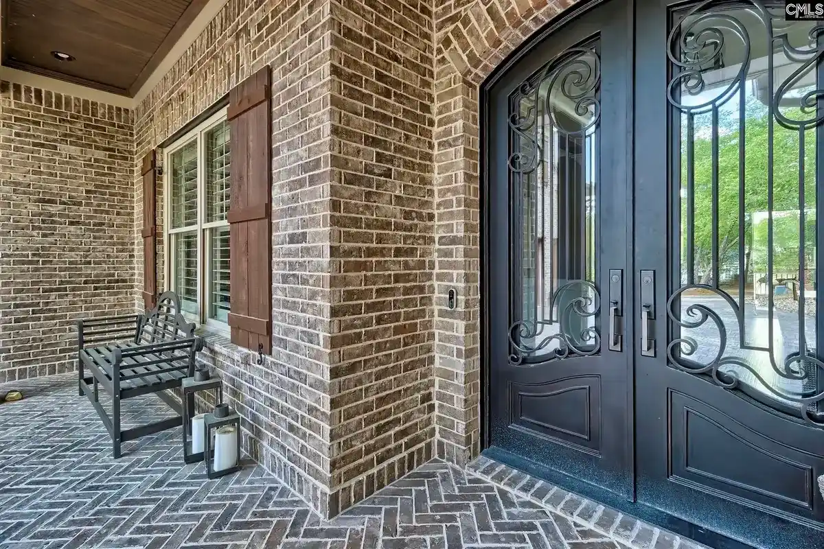 A brick porch with a herringbone pattern walkway leads to a grand double door entrance with ornate ironwork.