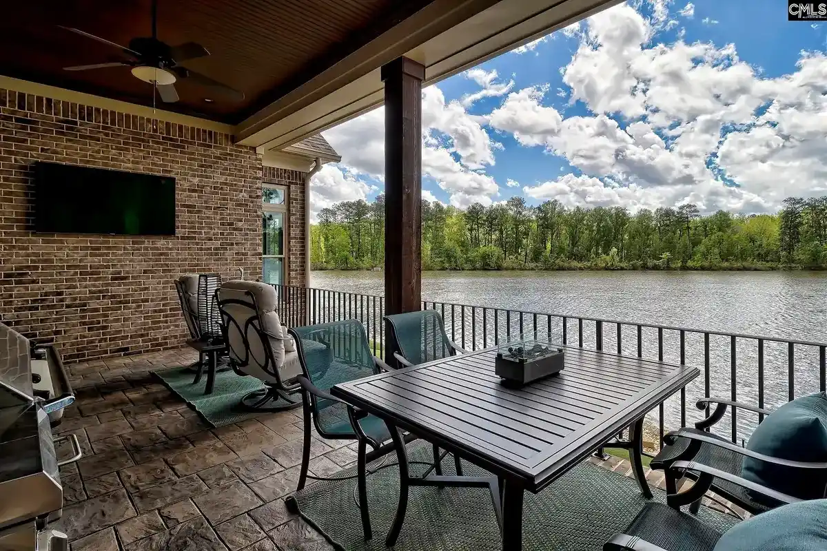 A brick patio with outdoor furniture and a flat-screen TV overlooks a calm lake under a partly cloudy sky.