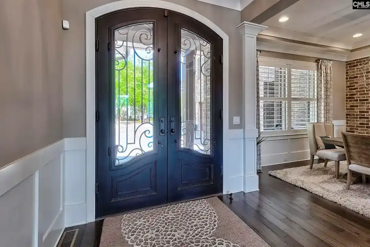 A grand entryway features double doors with intricate ironwork, a patterned rug, and hardwood floors, leading to a dining area with large windows.