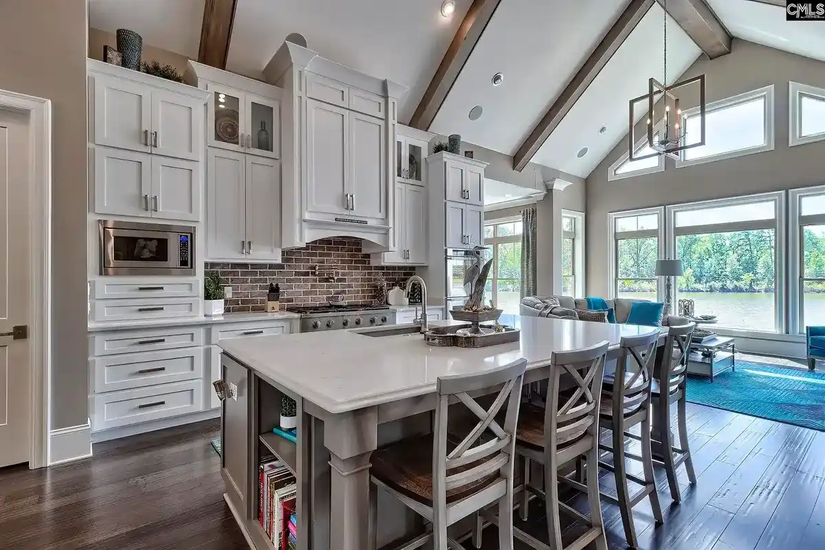 A kitchen with white cabinets, a large island, and a brick backsplash opens to a living area with large windows and a view of a lake.