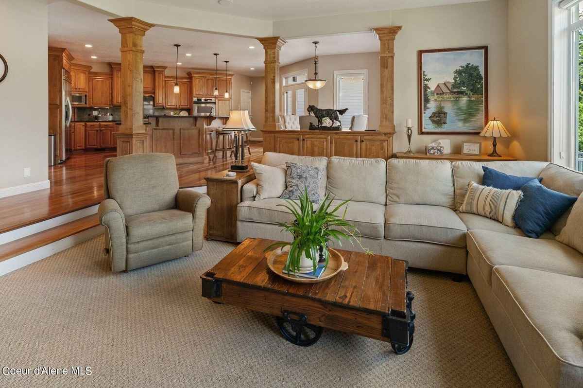 A large living room features a sectional sofa, a recliner, and a rustic coffee table, with a partially visible kitchen beyond wooden columns.