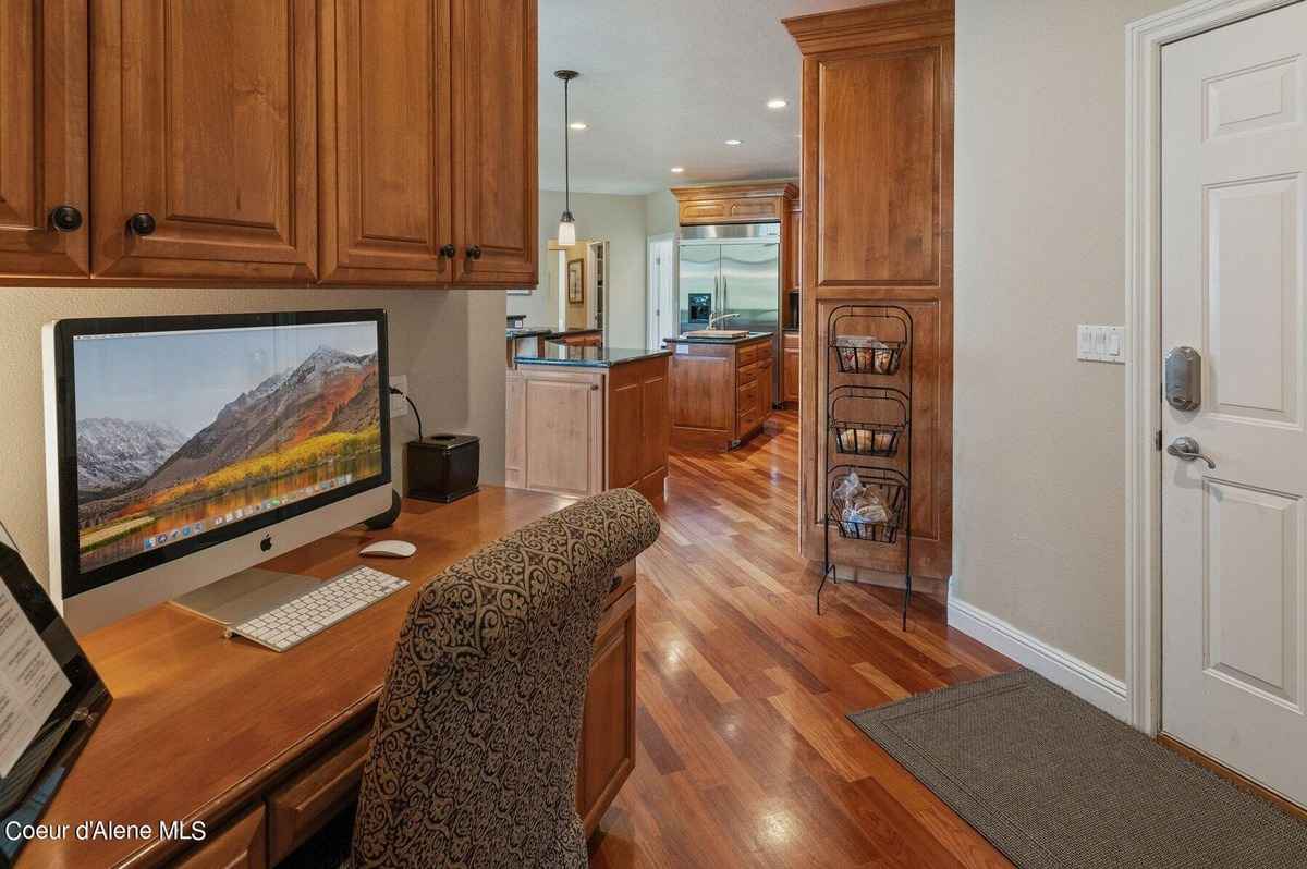 A home office with a computer, desk, and wood floors is shown. A partially visible kitchen is in the background.