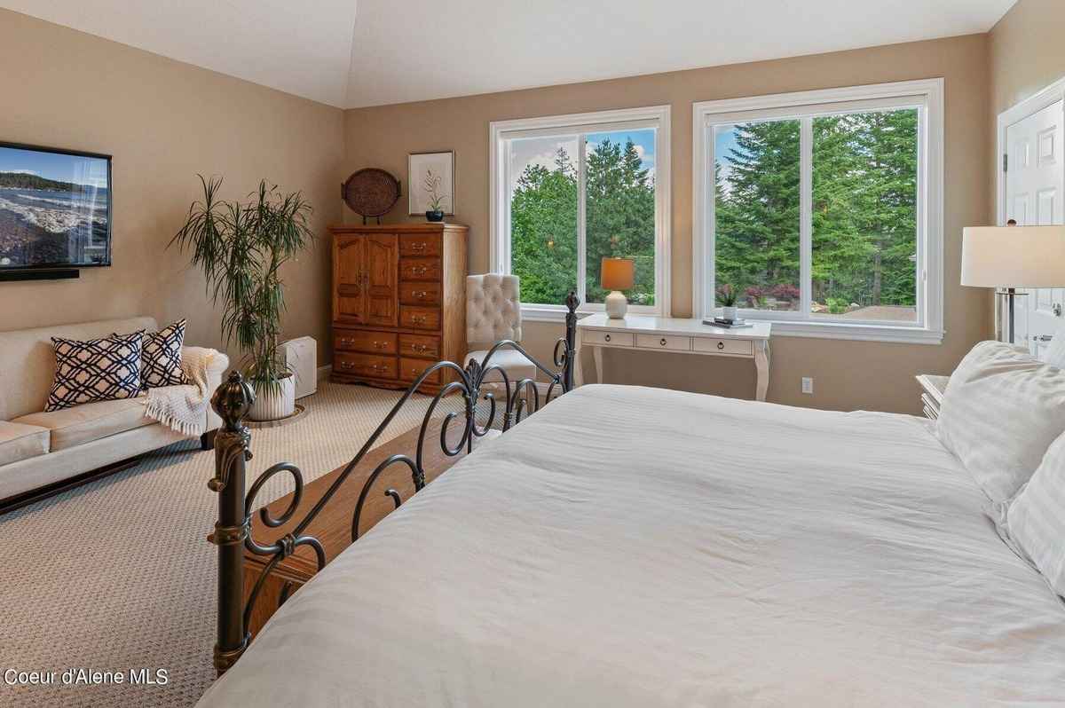 A bedroom with a large bed, a dresser, a plant, and a view of trees outside is shown.